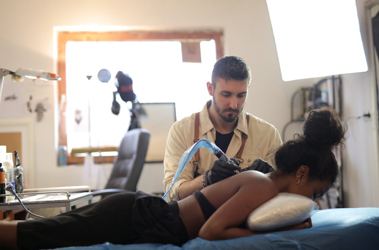 A tattoo artist concentrates as he inks a design on a woman's back in a bright studio setting.